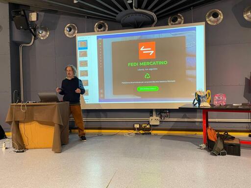 io sto in piedi di fronte a uno schermo con una presentazione.indosso una camicia nera e pantaloni blu. Ci sono degli altoparlanti in entrambi i lati dello schermo. Sul lato destro dello schermo c'è un tavolo con dei libri e dei cavi. Sul lato sinistro dello schermo ci sono delle casse. In cima allo schermo è presente la scritta "FEDI MERCATINO". L'ambiente è una sala conferenze.

