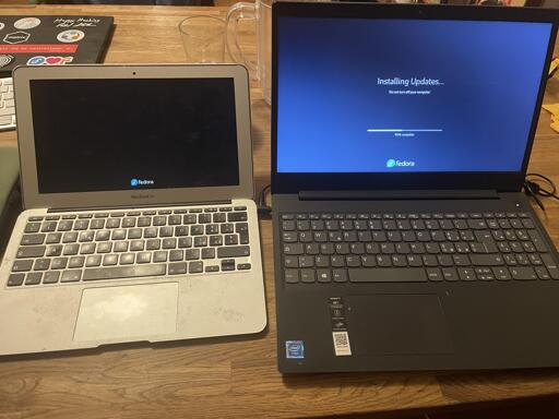 Due computer portatili sono affiancati su una superficie di legno. Il laptop a sinistra è argento con lo schermo nero che mostra il logo bianco "fedora" e la scritta "MacBook Air" sotto lo schermo. Il laptop a destra è grigio scuro con uno schermo blu che riporta il testo "Installing Updates.. Updates... Do not turn your computer off 40% complete" e il logo "fedora" in basso. In alto a sinistra si vede un quaderno con adesivi che riportano "matria" e "Happy Hacking!". Sul laptop di destra sono presenti un logo Intel e un piccolo adesivo con un codice QR.
