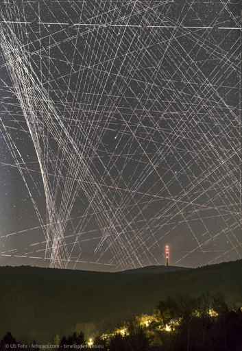 Una fotografia notturna a lunga esposizione mostra un cielo scuro attraversato da numerose scie luminose bianche di varie dimensioni e direzioni. Le scie si intrecciano in una rete complessa sopra piccoli punti luminosi. In basso, la silhouette scura di una collina delimita l'ori l'orizzonte, con una torre sottile che emette luci rosse e bianche su una delle vette. Nella parte inferiore si intravedono luci calde tra la vegetazione scura. In fondo all'immagine compare il testo: "© Uli Fehr - fehrpics.com - timelapsevideo.eu".