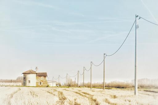Una cappella tra le stoppie di una risaia a fine inverno, alla sua destra corre una fila di pali che sorregge la linea telefonica.
Foto a colori naturali desaturati.