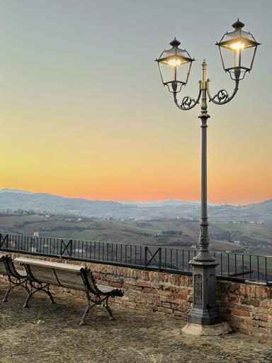 A vintage streetlamp stands beside two benches overlooking rolling hills at sunset, with a gradient sky transitioning from orange to blue. A stone wall encloses the area.