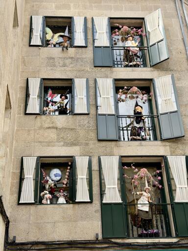 Figurine di cartapesta dipinte, raffiguranti personaggi fantastici, in balconi decorati con fiori, su una facciata di edificio di pietra.