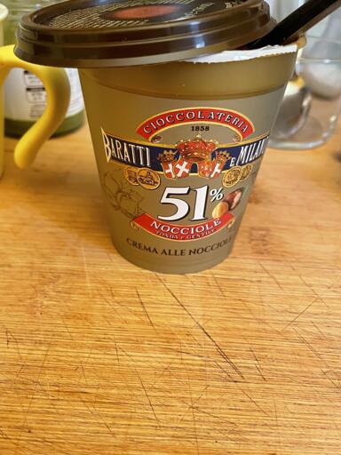 L'immagine mostra una vaschetta di crema alla nocciola posizionata su un tagliere di legno. La vaschetta è contenuta in una scatola bianca e marrone con un coperchio trasparente. Sulla scatola si legge "CIOCCOLATERIA MILANO". Sotto questa scritta, si legge "51%". Sotto, si legge "NOCCIOLE". Sotto queste parole, c'è un logo con il disegno stilizzato di una testa. Sotto a questo, si legge "1858". L'intero design è in tonalità di marrone, bianco e oro. Il tagliere di legno ha una grana visibile ed è leggermente sgualcito.