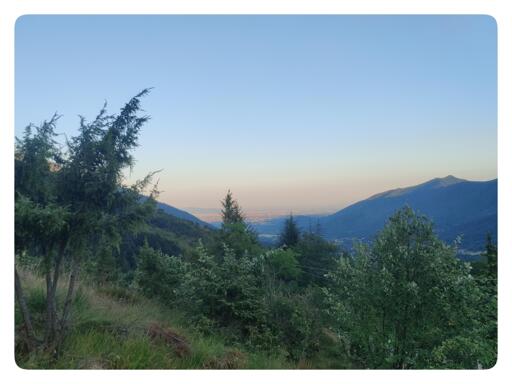 Paesaggio montano.
In primo piano, i bassi arbusti che ricoprono la riva. In lontananza i profili delle montagne più lontane. All'orizzonte si apre la pianura, con la foschia dovuta all'afa e il cielo arancio del tramonto.