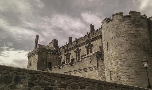 Fotografia che ci mostra il castello di Stirling sotto un cielo nuvoloso. L'inquadratura dal basso sottolinea la poderosa torre rotonda e le merlature che sovrastano il cortile, ma mostra anche le quattro nicchie con statue sulla facciata, che ne attestano la ricchezza delle decorazioni