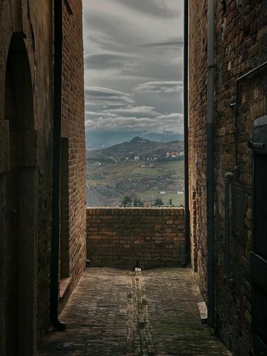 A narrow alleyway between brick buildings, leading to a scenic view of hills and cloudy skies in the distance.