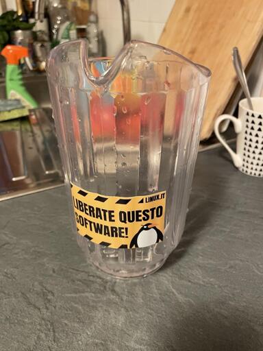 L'immagine mostra una caraffa trasparente in plastica posizionata su una superficie grigia, probabilmente una cucina. La caraffa ha un'etichetta gialla con bordi neri che recita "LIBERATE QUESTO SOFTWARE!" e "LINUX.IT", con un pinguino al centro. La caraffa ha condensa, suggerendo che è stata recentemente riempita con un liquido freddo. Sullo sfondo, si intravede un lavello con un rubinetto, un detergente verde e un asciugamano. A destra, c'è un bicchiere bianco con un motivo di triangoli neri. L'atmosfera è domestica e informale, con un tocco di humor e cultura open source.