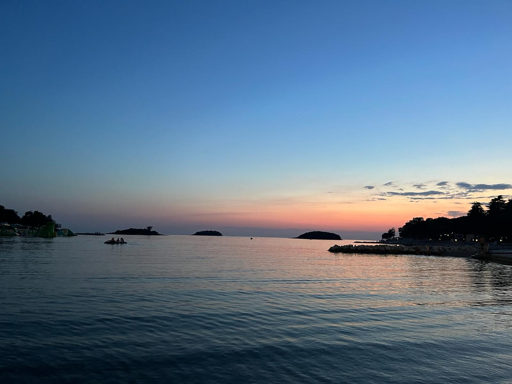 Una fotografia di un paesaggio marino con cielo e mare. Il cielo sfuma da un blu scuro nella parte superiore a una tonalità arancione chiaro all'oriz all'orizzonte, con un piccolo punto bianco visibile in alto al centro. Diverse sagome scure di isole e terraferma si stagliano lungo la linea dell'orizzonte. Sulla destra, una costa alberata presenta un lungo molo che si protende verso l'acqua. L'acqua calma in primo piano riflette i gradienti di colore del cielo.