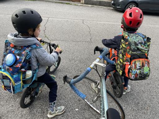 Due bambini sono in piedi sulle loro biciclette su una superficie di asfalto grigia. Il bambino a sinistra indossa una giacca grigia, pantaloni neri, scarpe grigie e un casco nero. Il bambino a destra indossa una maglietta scura, pantaloni scuri e un casco rosso. Entrambi i bambini stanno guardando l'uno nell'altro. Entrambi i bambini hanno uno zaino sulla schiena. Il bambino a sinistra ha uno zaino blu con immagini di personaggi, il bambino a destra ha uno zaino verde e giallo con immagini di draghi. Entrambi i bambini sono su biciclette con manubri neri e pedali. Il bambino a destra ha una fascia blu intorno al manubrio.
